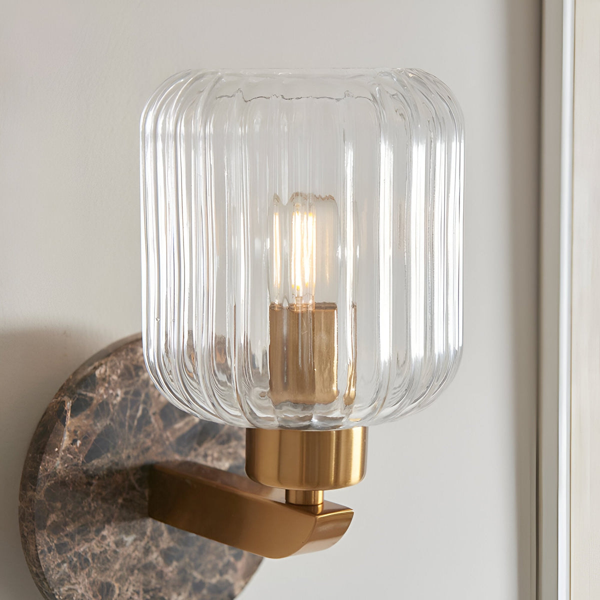 Cranton Wall Light in Brass & Brown featuring a ribbed clear glass shade on a brass arm, mounted on a dark Emperador marble backplate with visible veining.