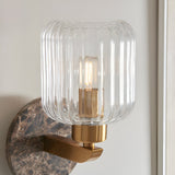 Cranton Wall Light in Brass & Brown featuring a ribbed clear glass shade on a brass arm, mounted on a dark Emperador marble backplate with visible veining.