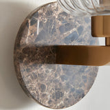 Cranton Wall Light in brass, close-up of the round Emperador marble backplate with brown veining, brass arm, and ribbed clear glass shade.