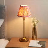Endon Cliffbourne Ikat table lamp with stepped brushed brass base and pleated pink Ikat faux silk shade, lit on a bedside table beside a brass vase.