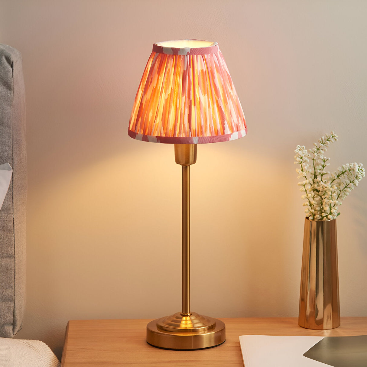 Endon Cliffbourne Ikat table lamp with brushed brass stepped base and pleated pink Ikat faux silk shade, lit on a bedside table beside a brass vase.