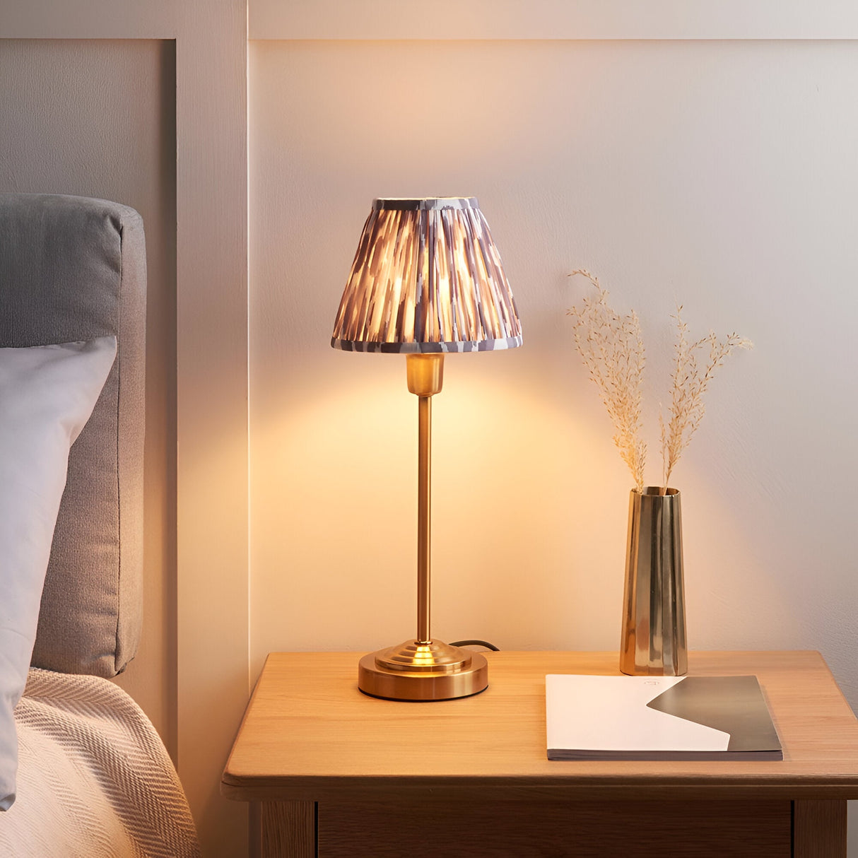 Cliffbourne Ikat Table Lamp with brass stepped base and pleated grey Ikat shade, glowing on a wooden bedside table.
