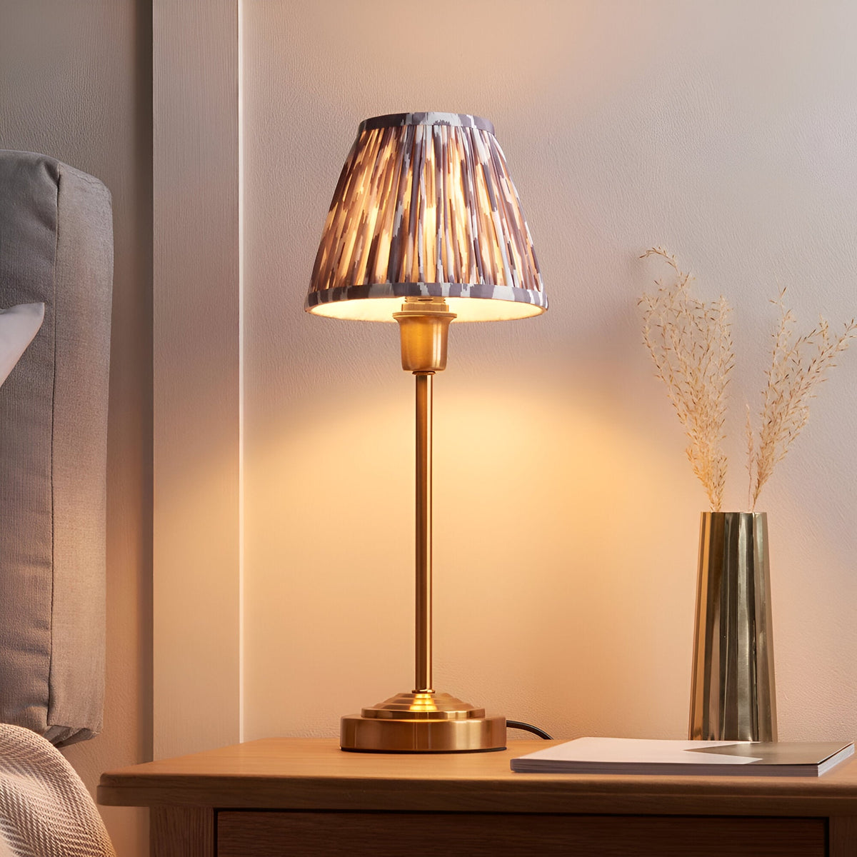 Cliffbourne Ikat Table Lamp with a brass stepped base and pleated grey Ikat fabric shade, shown lit on a bedside table.