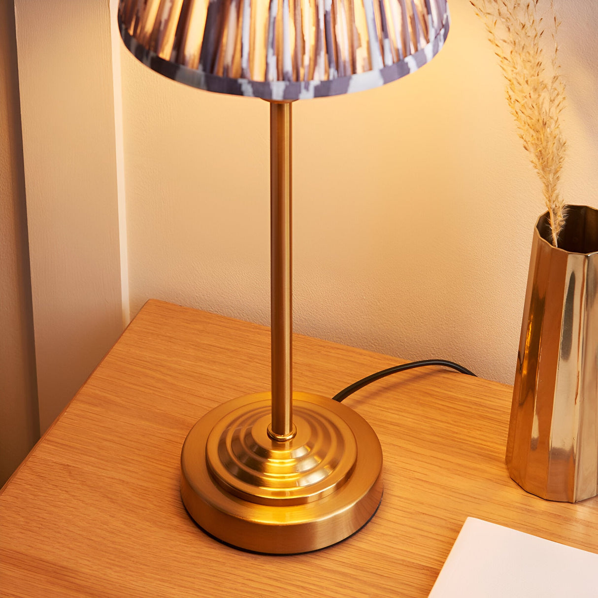 Cliffbourne Ikat table lamp with a stepped brass base and pleated grey Ikat fabric shade, shown lit on a wooden bedside table.