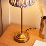 Cliffbourne Ikat table lamp with a stepped brass base and pleated grey Ikat fabric shade, shown lit on a wooden bedside table.