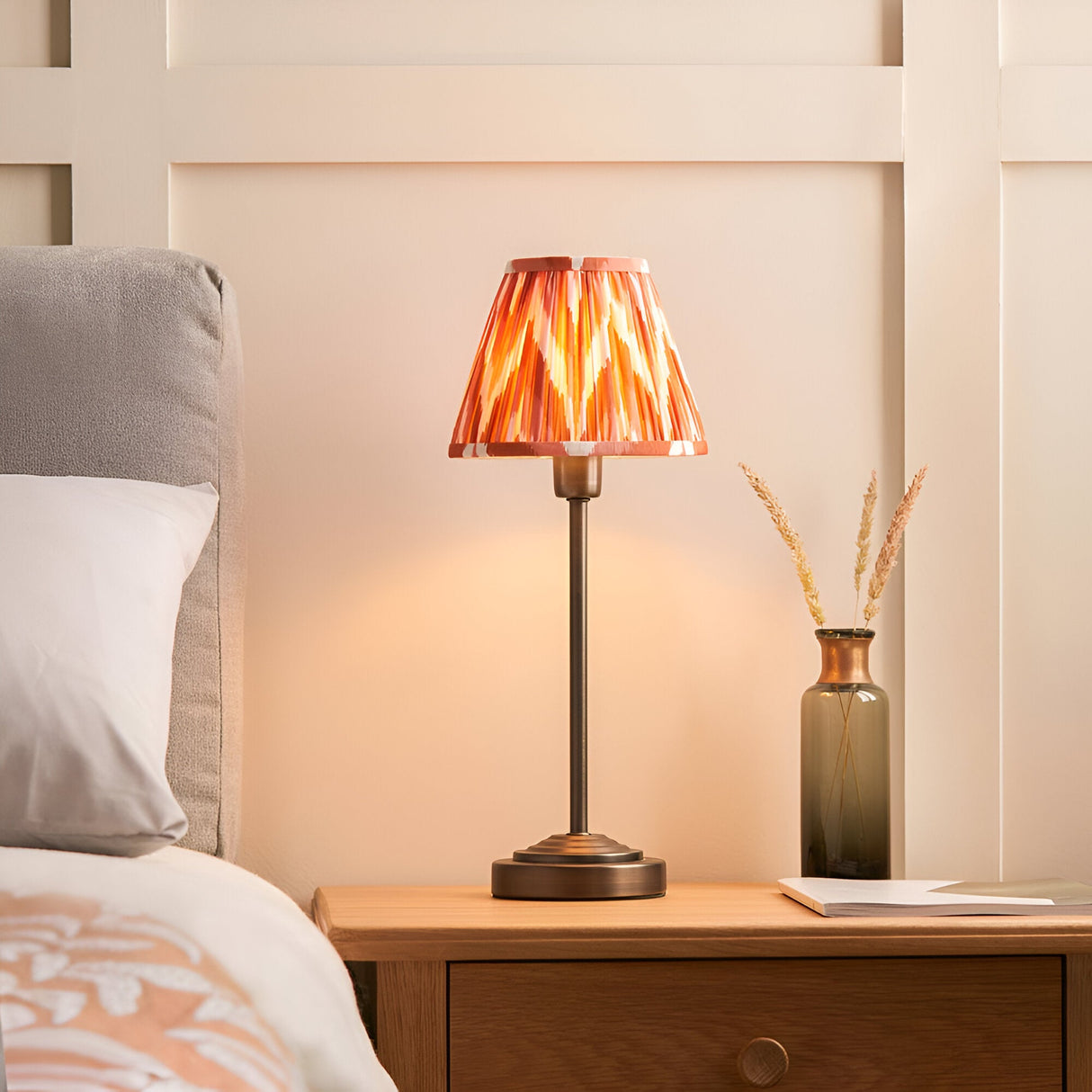 Endon Cliffbourne Zigzag table lamp with bronze stepped base and tapered pleated apricot zigzag shade, on a bedside table beside a bed and dried grass vase.