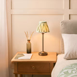 Endon Cliffbourne Zigzag table lamp with bronze stepped base and green pleated faux silk shade, lit on a wooden bedside table beside a vase in a bedroom.