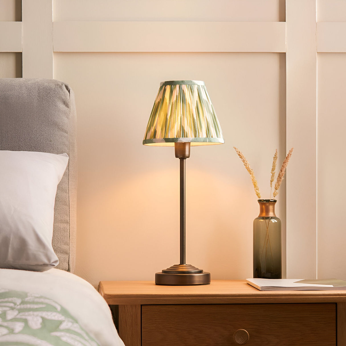 Endon Cliffbourne Zigzag table lamp with bronze stepped base and green pleated faux silk shade, lit on a wooden bedside table beside a glass vase.