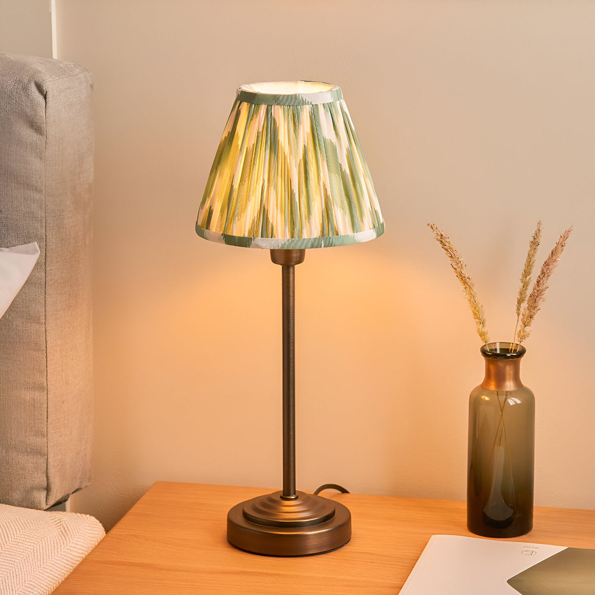 Endon Cliffbourne Zigzag table lamp with antique bronze stepped base and tapered pleated green zigzag shade, lit on a bedside table beside a glass vase.