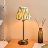 Endon Cliffbourne Zigzag table lamp with antique bronze stepped base and tapered pleated green zigzag shade, lit on a bedside table beside a glass vase.
