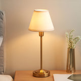 Cliffbourne Ivy Table Lamp with brass stepped base and tapered white fabric shade, shown lit on a wooden bedside table beside a vase.
