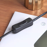 Close-up of a black inline cord switch on a lamp cable resting on a white notebook, shown on a wooden desk with a brass accent object.