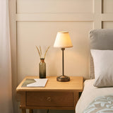 Endon Cliffbourne Ivy table lamp with stepped antique bronze base and tapered vintage white shade, lit on an oak bedside drawer beside a bed.