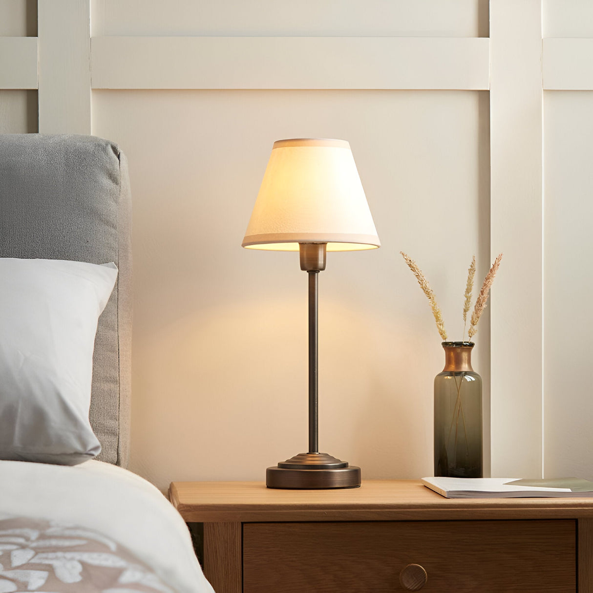 Endon Cliffbourne Ivy table lamp with slim antique bronze stepped base and tapered vintage white shade, lit on a wooden bedside table beside a bed.