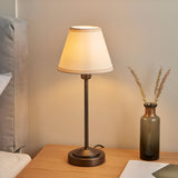 Endon Cliffbourne Ivy table lamp with stepped antique bronze base and tapered vintage white fabric shade, lit on a wooden bedside table beside a glass vase.