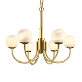 The Aurelio 6 Light Large Globe Alabaster Chandelier features a brushed brass frame with six curved arms, each holding a soft white alabaster globe, suspended from a matching chain for a modern look.