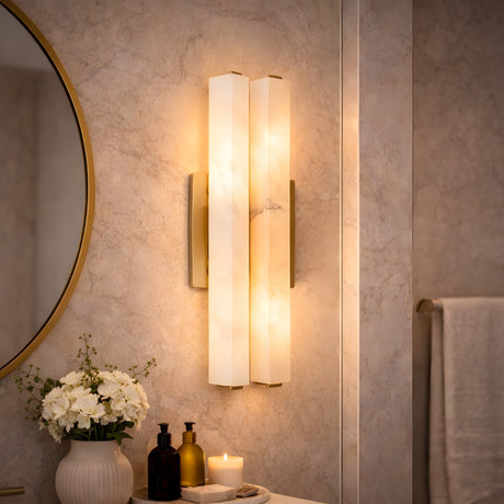 A modern bathroom with the Selvara LED Medium Alabaster Double Wall Light in Brushed Brass & Soft White, glowing above decorative bottles, a candle, and a vase of white flowers on a countertop beneath a round mirror. Cream walls and towel in back.