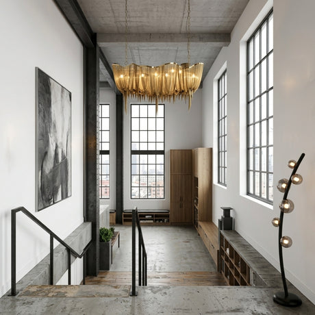 A modern loft with tall windows, concrete floor, gold chandelier, abstract art, and minimalist furniture. Natural light brightens the open staircase and décor, complemented by the Cordaro 6 Light Leather Floor Lamp - Black for sophistication.