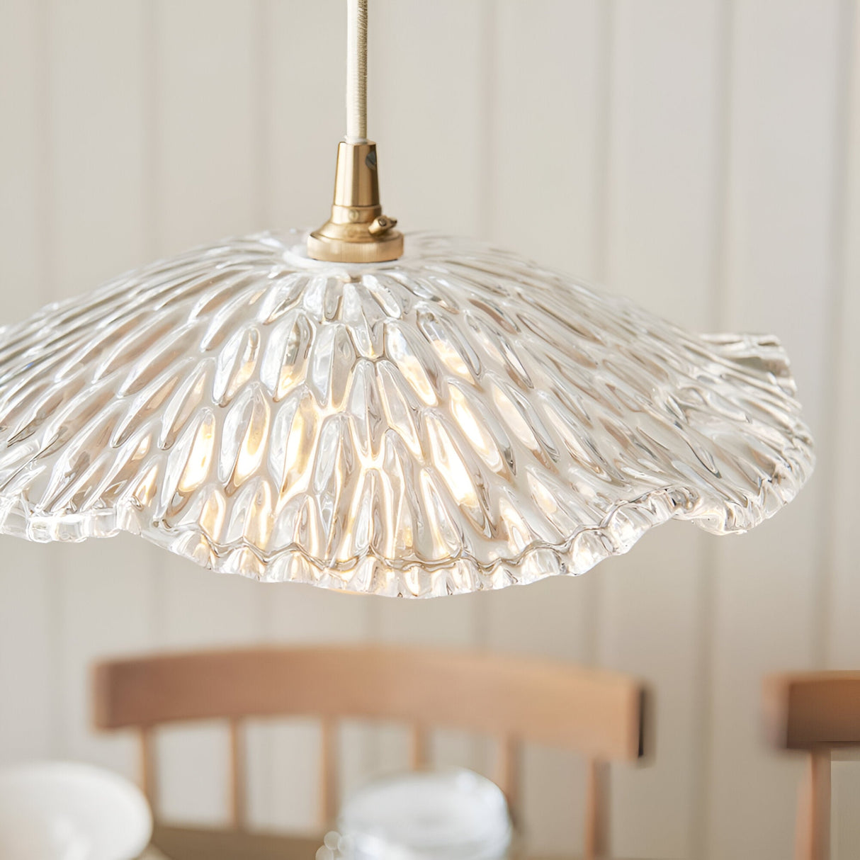 Brackmont Orchid Pendant in Brass & Natural, featuring a rippled clear glass shade on a brass fitting, shown lit above a dining area with wooden chairs.