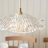 Brackmont Orchid Pendant in Brass & Natural, featuring a rippled clear glass shade on a brass fitting, shown lit above a dining area with wooden chairs.