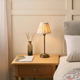 Cliffbourne Ikat Table Lamp in bronze with a stepped metal base and pleated fabric shade, shown lit on a wooden bedside table beside a bed.