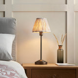 Cliffbourne Ikat Table Lamp in bronze with a stepped metal base and pleated fabric shade, shown lit on a bedside table.