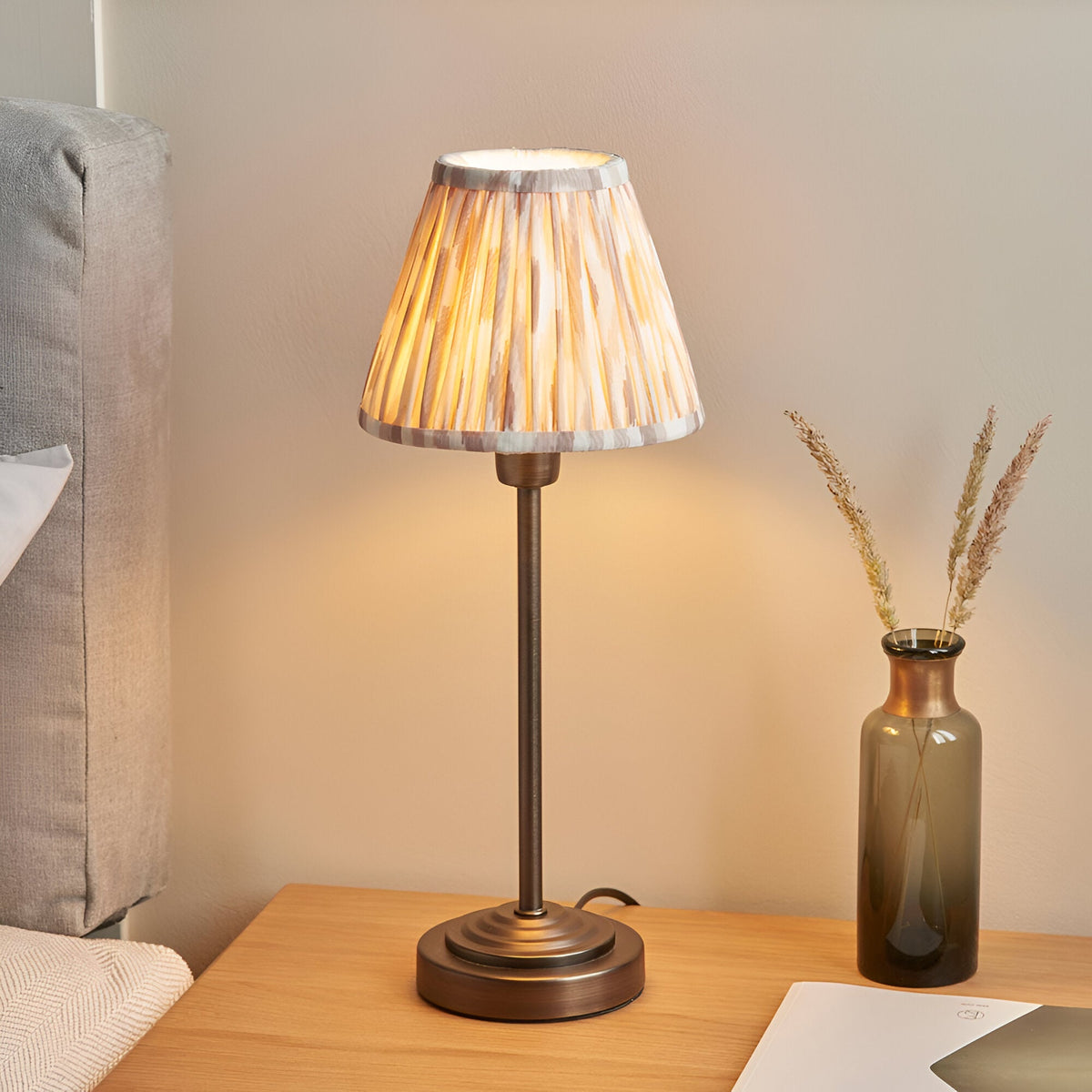 Cliffbourne Ikat Table Lamp in bronze with a stepped metal base and pleated Ikat-print fabric shade, shown lit on a bedside table.
