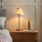 Endon Cliffbourne Ikat table lamp with brushed brass stepped base and pleated Ikat shade, lit on a wooden bedside table beside a bed and vase.