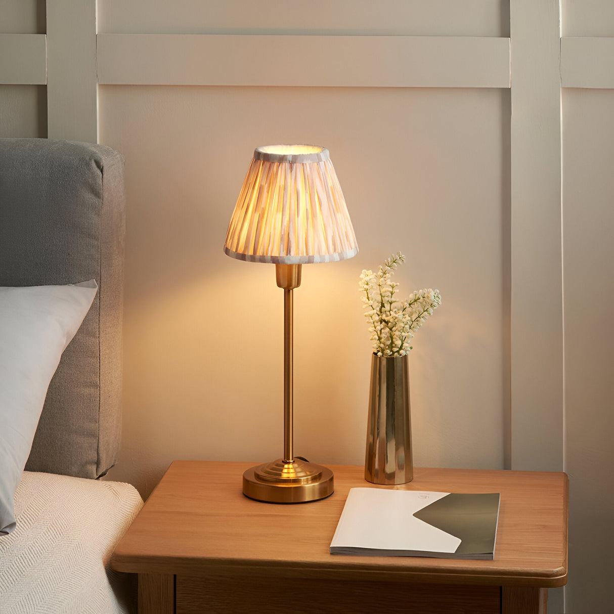 Endon Cliffbourne Ikat table lamp with brass stepped base and pleated printed faux silk shade, lit on a wooden bedside table beside a brass vase.