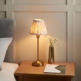 Endon Cliffbourne Ikat table lamp with brass stepped base and pleated printed faux silk shade, lit on a wooden bedside table beside a brass vase.
