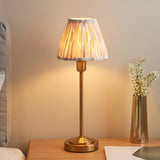 Endon Cliffbourne Ikat table lamp with brushed brass stepped base and pleated grey Ikat-print faux silk shade, lit on a bedside table beside dried flowers.