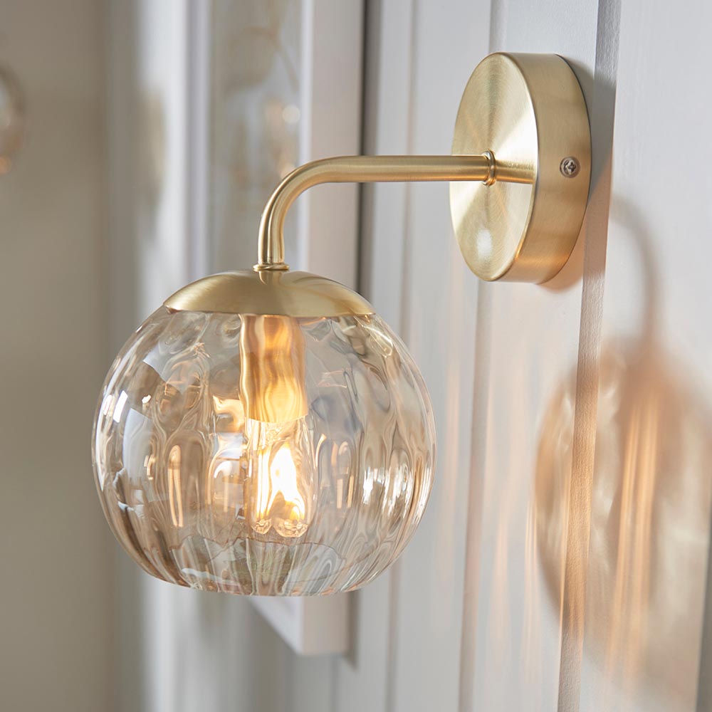 Endon Draycott wall light in satin brass with a champagne lustre dimpled glass globe shade, mounted on a white panelled wall with the lamp lit.