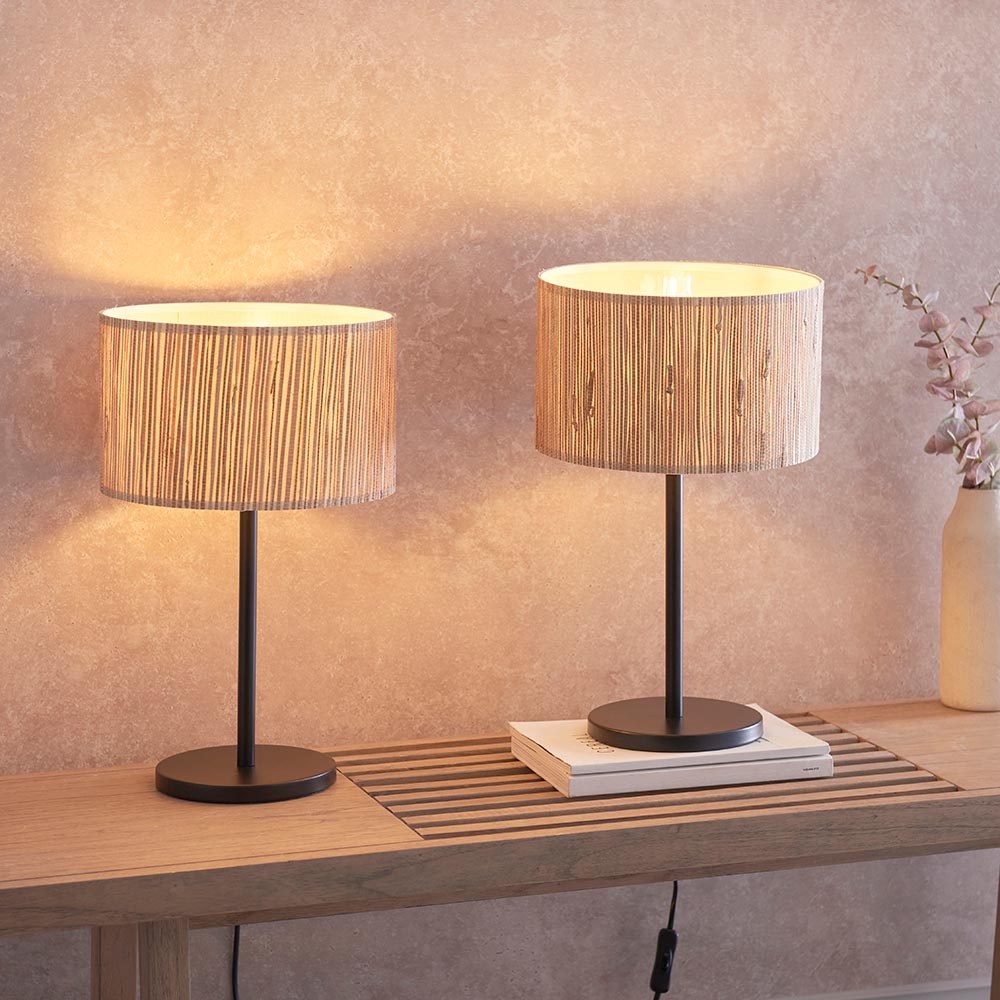 Salcombe Table Lamp Twin Pack in black, two lit lamps with woven seagrass drum shades on slim metal stands, shown on a wooden console table.