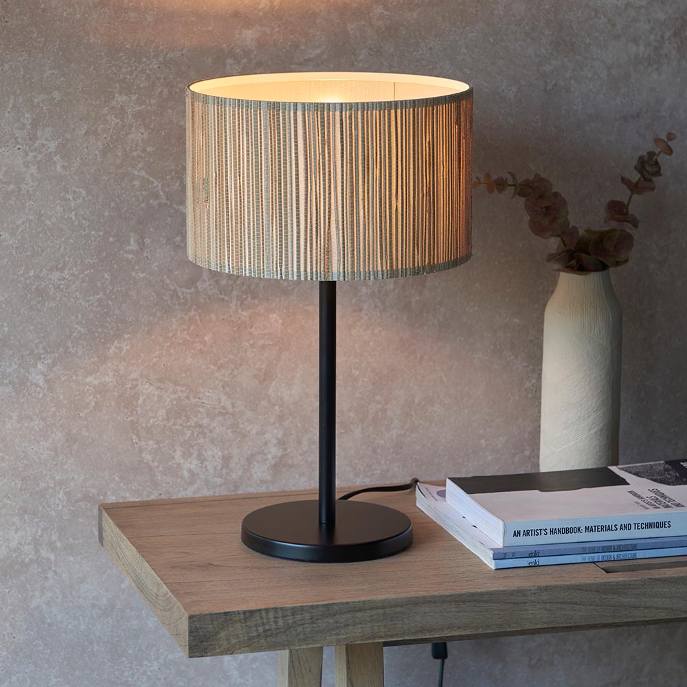 Salcombe Table Lamp with matt black base and stem, woven seagrass drum shade, lit on a wooden console table beside books and a vase.