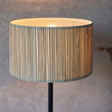 Salcombe Table Lamp with woven seagrass drum shade on a matt black metal base, glowing warmly against a neutral backdrop.