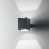 The Opus Square Outdoor Wall Light in anthracite, with its modern cube shape, is mounted on a white wall. It casts light both upward and downward through Pyrex glass, creating a geometric pattern of light and shadows.