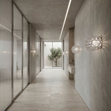 Minimalist hallway with light gray walls and floor, glass partitions on the left, Briller LED Large Wall Light - Transparent installed, a simple sink on the right, and a large window at the end revealing a small tree outside.