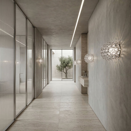Minimalist hallway with light gray walls and floor, glass partitions on the left, Briller LED Large Wall Light - Transparent installed, a simple sink on the right, and a large window at the end revealing a small tree outside.