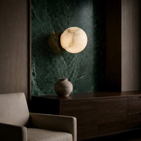 A modern interior features a cream armchair, a dark wooden sideboard with a ceramic vase, and the Alviora 1 Light Small Circular Stone Alabaster Wall Light – Soft White & Black glowing against a green marble accent wall.