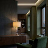 A modern living room with dark wood furniture, a green armchair, and the Noirion 1 Light Stacked Marble Table Lamp (Brass & White) atop a side table. Large windows reveal city buildings at dusk; warm, soft lighting and built-in shelves complete the scene.
