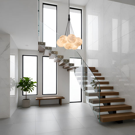 A modern, minimalist staircase with wooden steps and glass railings stands in a bright room with tall windows, a potted plant, a wooden bench, and the Auriel 7 Light Alabaster Chandelier - Soft White & Black hanging above.