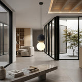 A modern minimalist living space with floor-to-ceiling glass walls, a round white armchair, built-in shelves, an open courtyard tree, and the Origo Concrete Pendant Light - Black & White featuring contrasting spheres.