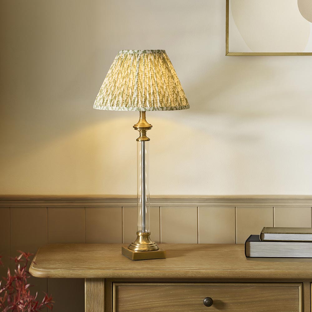 Endon Aldmont Leaf table lamp with a clear glass column, antique brass base and cap, and a pleated green leaf-print shade on a wooden sideboard.