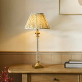 Endon Aldmont Leaf table lamp with a clear glass column, antique brass base and cap, and a pleated green leaf-print shade on a wooden sideboard.