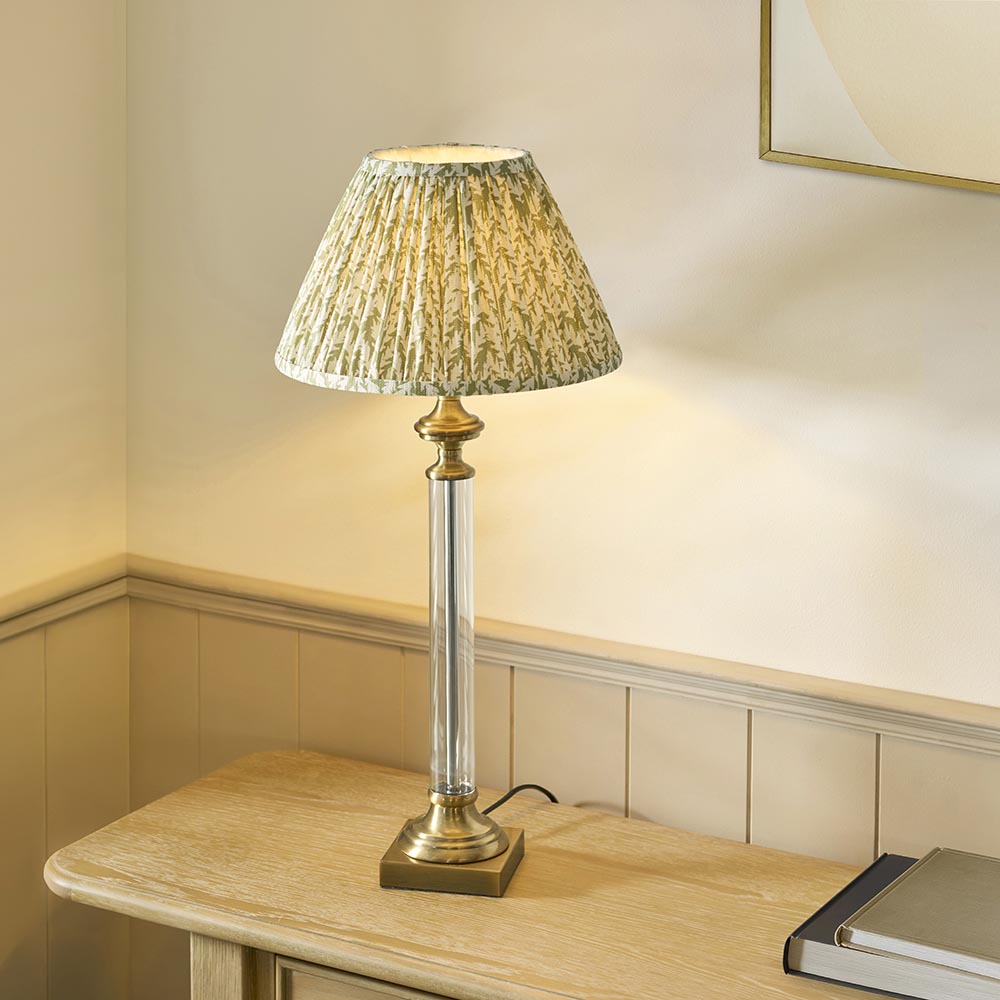 Endon Aldmont Leaf table lamp lit on a wooden sideboard, with clear glass column, antique brass base and pleated green leaf-print fabric shade.