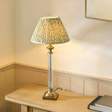 Endon Aldmont Leaf table lamp lit on a wooden sideboard, with clear glass column, antique brass base and pleated green leaf-print fabric shade.