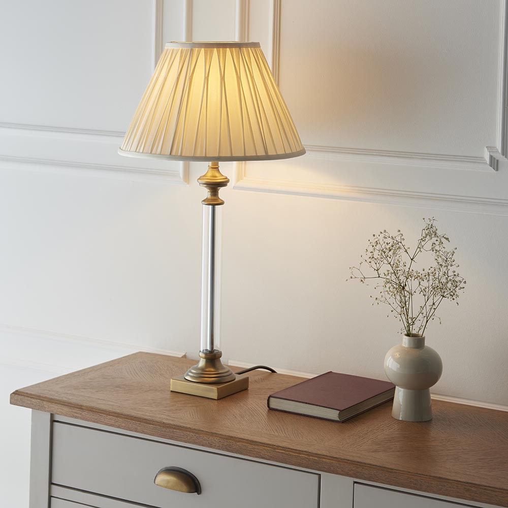 Endon Aldmont Leaf table lamp with clear glass column, antique brass base and pleated cream shade on a wooden sideboard beside a book and dried flowers.