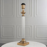 Endon Aldmont table lamp base with antique brass cap, clear glass column and square brass foot, shown on a marble table against panelled wall.