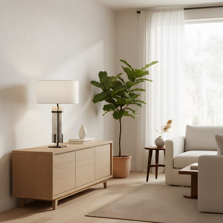 A bright, modern living room with light wood furniture, a potted plant, a Pilaster 1 Light Column Alabaster Table Lamp in matte black and soft white on the sideboard, a small round table, white sofa, and sheer curtains letting in natural light.