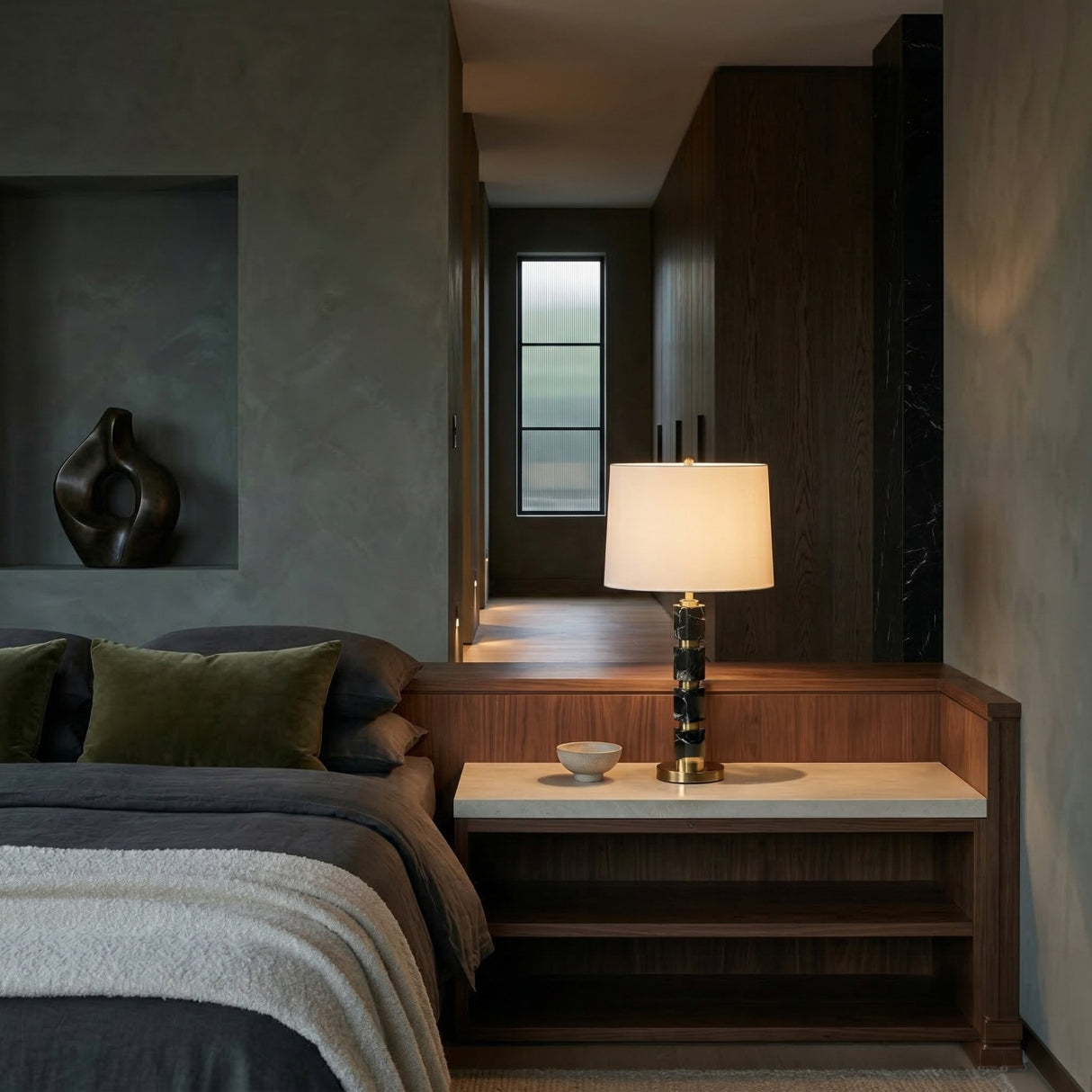 A modern bedroom with a dark bed, green pillows, a wood headboard with built-in side table, and the Noirion 1 Light Stacked Marble Table Lamp in brass & black. A small bowl and sculpture are on the shelf, with a windowed hallway in back.
